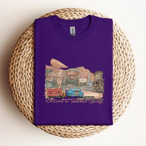 May include: A purple t-shirt featuring a colorful illustration of a desert town with vintage cars and the text "Welcome to Radiator Springs". The design includes a red truck, a blue car, and a water tower.