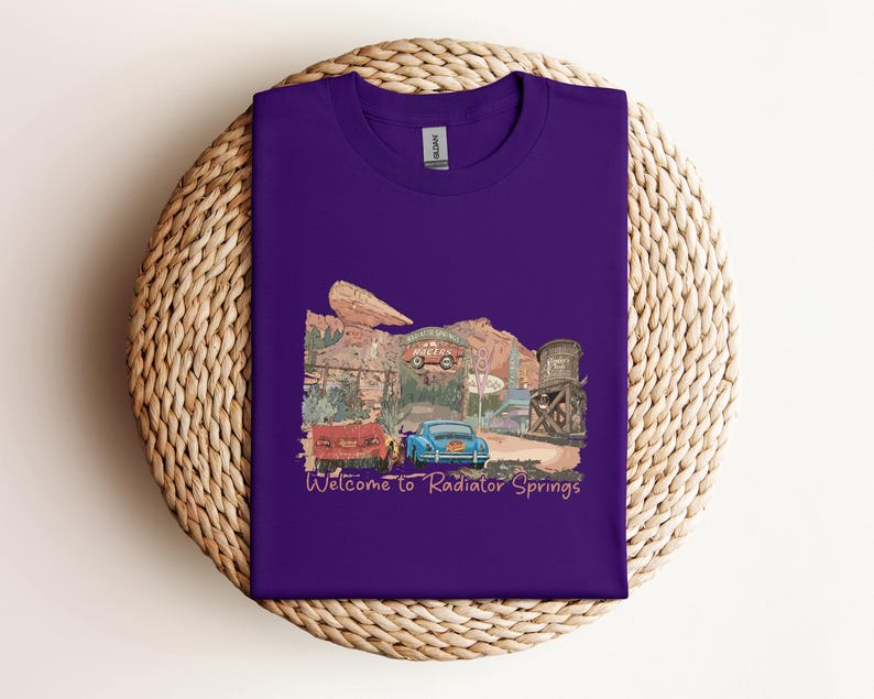 May include: A purple t-shirt featuring a colorful illustration of a desert town with vintage cars and the text "Welcome to Radiator Springs". The design includes a red truck, a blue car, and a water tower.