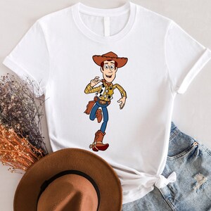 May include: White t-shirt with a cartoon image of Woody from Toy Story. Woody is wearing his signature cowboy hat, vest, and boots. He is holding his arm out and smiling.