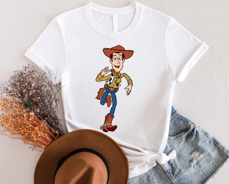 May include: White t-shirt with a cartoon image of Woody from Toy Story. Woody is wearing his signature cowboy hat, vest, and boots. He is holding his arm out and smiling.