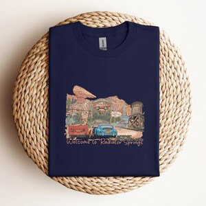 May include: Navy blue t-shirt featuring a colorful illustration of a desert town with vintage cars and the text "Welcome to Radiator Springs." The design includes a red car, a blue car, and a water tower. The shirt is made of a soft material.