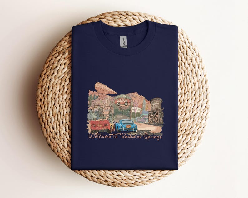 May include: Navy blue t-shirt featuring a colorful illustration of a desert town with vintage cars and the text "Welcome to Radiator Springs." The design includes a red car, a blue car, and a water tower. The shirt is made of a soft material.