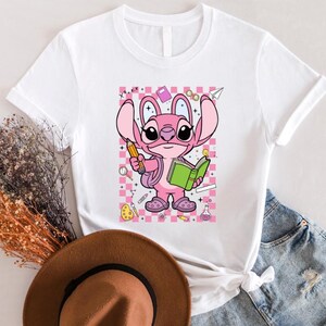 Puede incluir: Camiseta blanca con un personaje de dibujos animados rosa sosteniendo un l&aacute;piz y un libro verde. El personaje est&aacute; sobre un fondo a cuadros rosa y blanco con gr&aacute;ficos escolares. La camiseta tiene mangas cortas.