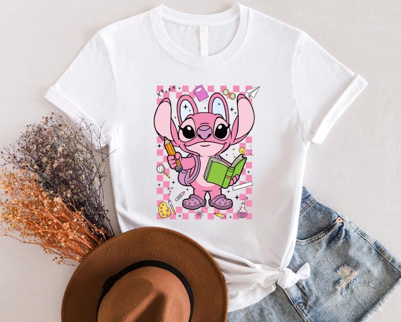 Puede incluir: Camiseta blanca con un personaje de dibujos animados rosa sosteniendo un l&aacute;piz y un libro verde. El personaje est&aacute; sobre un fondo a cuadros rosa y blanco con gr&aacute;ficos escolares. La camiseta tiene mangas cortas.
