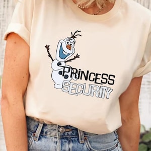以下が含まれることがあります： クリーム色のTシャツ。漫画の雪だるまのキャラクターと、黒と青で「PRINCESS SECURITY」という文字がプリントされています。雪だるまは笑顔で、腕を広げています。