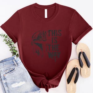 May include: A maroon t-shirt featuring a black graphic of a helmet and the text "THIS IS THE WAY". The shirt is paired with light blue denim shorts and beige flip-flops with black straps.