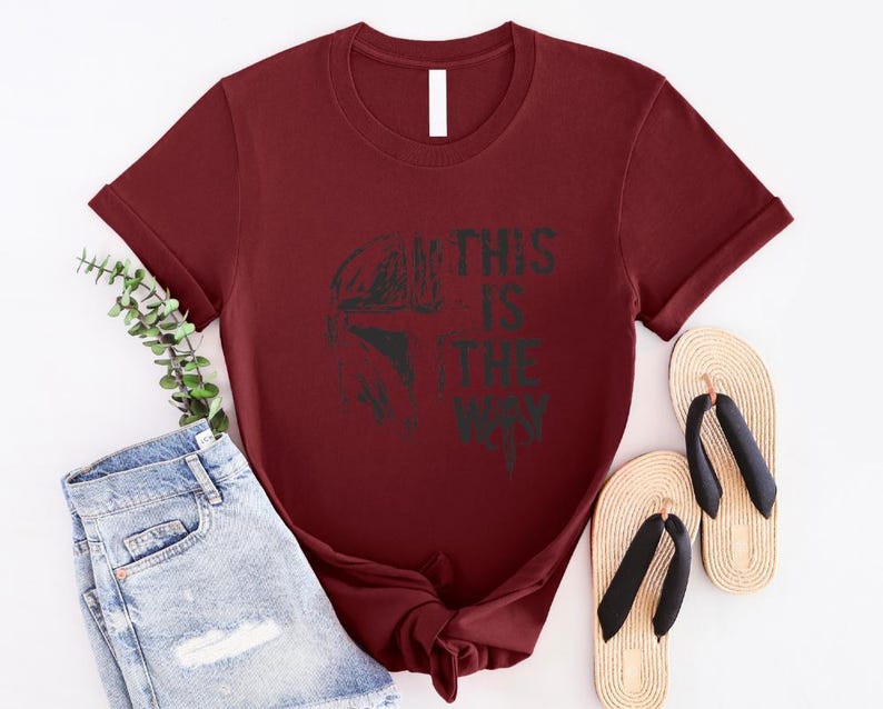 May include: A maroon t-shirt featuring a black graphic of a helmet and the text "THIS IS THE WAY". The shirt is paired with light blue denim shorts and beige flip-flops with black straps.