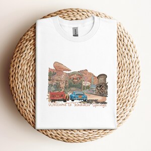 May include: White t-shirt featuring a watercolor illustration of Radiator Springs with two cars and the text "Welcome to Radiator Springs." The design includes a red car, a blue car, and a water tower.