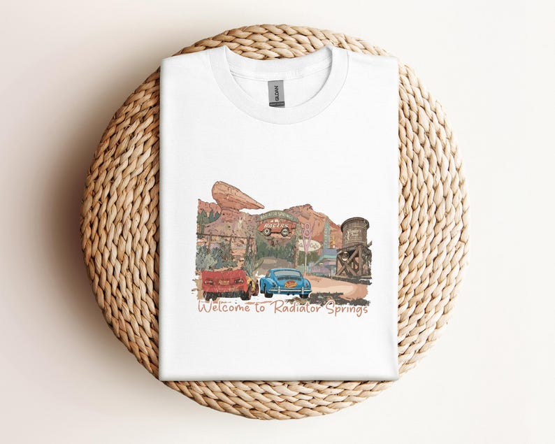 May include: White t-shirt featuring a watercolor illustration of Radiator Springs with two cars and the text "Welcome to Radiator Springs." The design includes a red car, a blue car, and a water tower.