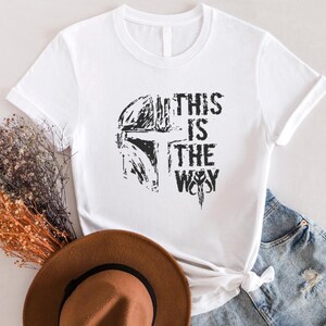May include: White t-shirt featuring a black graphic of a helmet and the text "THIS IS THE WAY." The design is distressed, giving it a vintage look. The shirt is styled with denim shorts and a brown hat.