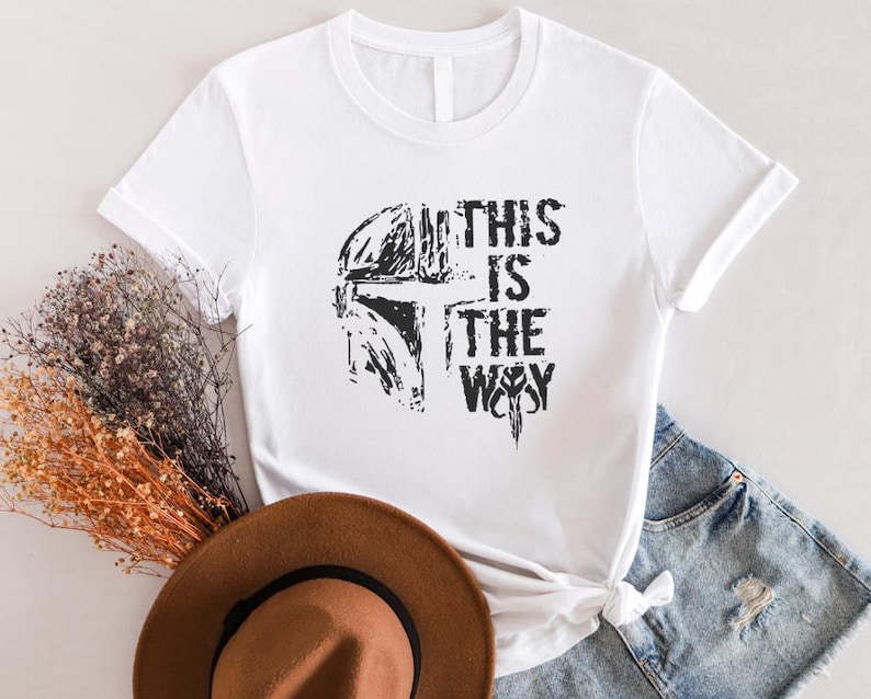 May include: White t-shirt featuring a black graphic of a helmet and the text "THIS IS THE WAY." The design is distressed, giving it a vintage look. The shirt is styled with denim shorts and a brown hat.