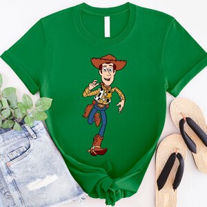 May include: A green t-shirt with a cartoon image of Woody from Toy Story. Woody is wearing his signature cowboy hat, vest, and boots. He is holding a sheriff's badge and a lasso.