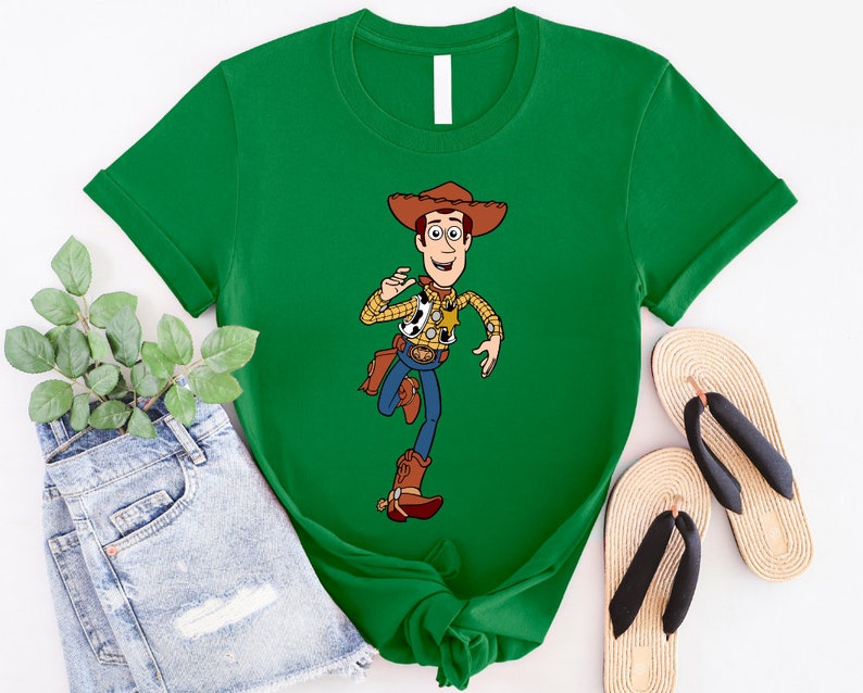 May include: A green t-shirt with a cartoon image of Woody from Toy Story. Woody is wearing his signature cowboy hat, vest, and boots. He is holding a sheriff's badge and a lasso.