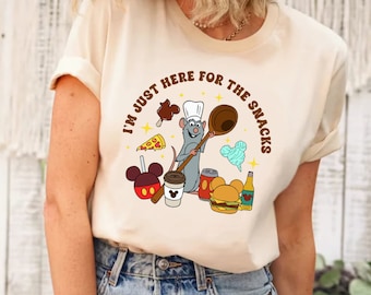 Può includere: Una maglietta bianca con una grafica marrone di un ratto chef che tiene un mestolo e circondato da vari snack, tra cui un gelato Mickey Mouse, una pizza, una mela Mickey Mouse, una tazza di caffè, una lattina di soda, zucchero filato e un hamburger. Il testo "I'm just here for the snacks" è scritto in un cerchio marrone attorno alla grafica.