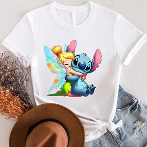 K&ouml;nnte beinhalten: Wei&szlig;es T-Shirt mit einem bunten Cartoon-Bild von Tinkerbell, die Stitch umarmt. Stitch ist blau mit einem rosa Ohr und einer rosa Nase. Tinkerbell hat blonde Haare und ein gr&uuml;nes Kleid.