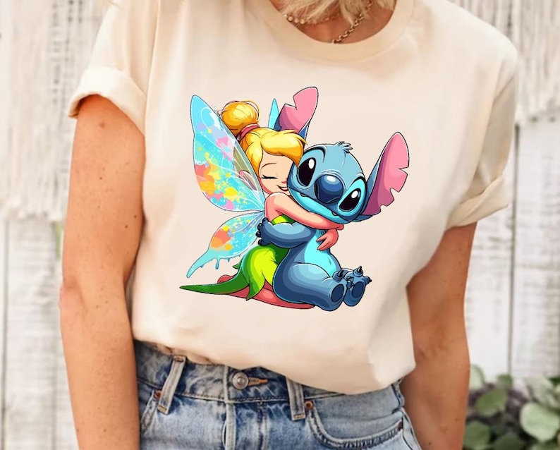 K&ouml;nnte beinhalten: Ein wei&szlig;es T-Shirt mit einem Cartoon-Bild von Tinkerbell, die Stitch umarmt. Tinkerbell hat gr&uuml;ne Fl&uuml;gel mit bunten Akzenten und Stitch ist blau mit rosa Ohren.