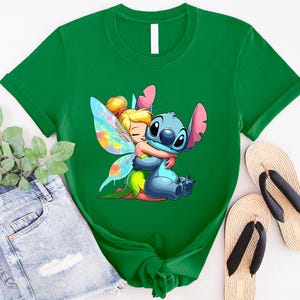 K&ouml;nnte beinhalten: Gr&uuml;nes T-Shirt mit einem Cartoon-Bild von Stitch aus Lilo & Stitch, der Tinkerbell aus Peter Pan umarmt. Stitch ist blau und Tinkerbell tr&auml;gt ein gr&uuml;nes Kleid mit Feenfl&uuml;geln.