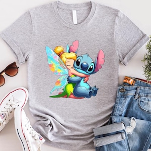 K&ouml;nnte beinhalten: Ein graues T-Shirt mit einem Cartoon-Bild von Stitch aus Lilo & Stitch, der Tinkerbell aus Peter Pan umarmt. Stitch ist blau und Tinkerbell tr&auml;gt ein gr&uuml;nes Kleid mit Feenfl&uuml;geln.