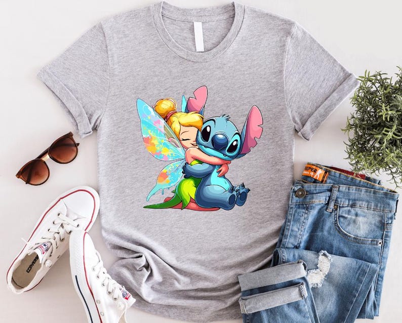 K&ouml;nnte beinhalten: Ein graues T-Shirt mit einem Cartoon-Bild von Stitch aus Lilo & Stitch, der Tinkerbell aus Peter Pan umarmt. Stitch ist blau und Tinkerbell tr&auml;gt ein gr&uuml;nes Kleid mit Feenfl&uuml;geln.