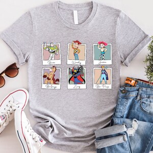 Puede incluir: Camiseta gris con un estampado de seis personajes de Toy Story en marcos Polaroid. Los personajes son Buzz Lightyear, Woody, Jessie, Bullseye, Zurg y Bo Peep. El texto debajo de cada marco dice "Buzz", "Woody", "Jessie", "Bullseye", "Zurg" y "Bo Peep".
