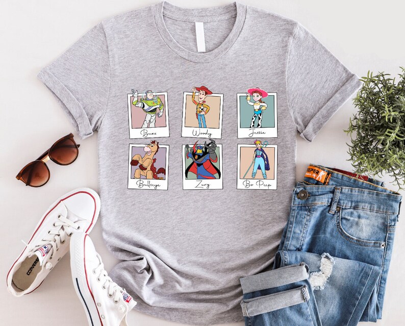 Puede incluir: Camiseta gris con un estampado de seis personajes de Toy Story en marcos Polaroid. Los personajes son Buzz Lightyear, Woody, Jessie, Bullseye, Zurg y Bo Peep. El texto debajo de cada marco dice "Buzz", "Woody", "Jessie", "Bullseye", "Zurg" y "Bo Peep".