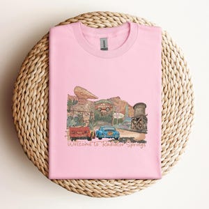 May include: A light pink t-shirt featuring a graphic of a desert town scene with two cars and the text "Welcome to Radiator Springs." The design includes a red truck, a blue car, and a water tower.