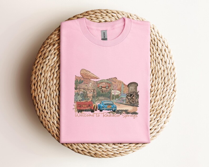May include: A light pink t-shirt featuring a graphic of a desert town scene with two cars and the text "Welcome to Radiator Springs." The design includes a red truck, a blue car, and a water tower.