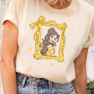 Pode incluir: Camiseta creme com uma ilustração de desenho animado de uma mulher em um vestido amarelo dentro de uma moldura dourada com um laço. Duas estrelas amarelas também estão presentes. A mulher tem cabelo castanho e está sorrindo.