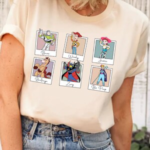 Puede incluir: Una camiseta de color crema con un gr&aacute;fico de seis personajes de Toy Story en marcos Polaroid. Los personajes incluyen Buzz Lightyear, Woody, Jessie, Bullseye, Zurg y Bo Peep.
