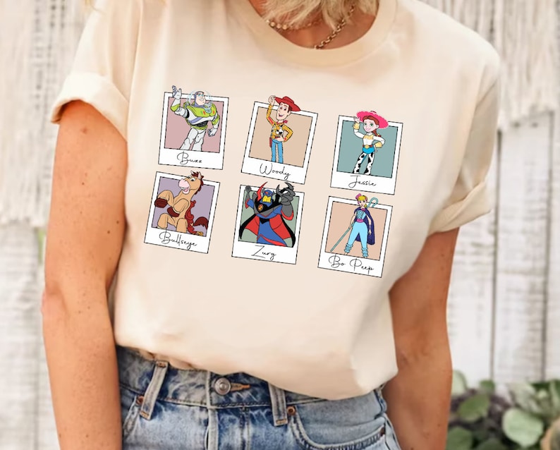 Puede incluir: Una camiseta de color crema con un gr&aacute;fico de seis personajes de Toy Story en marcos Polaroid. Los personajes incluyen Buzz Lightyear, Woody, Jessie, Bullseye, Zurg y Bo Peep.