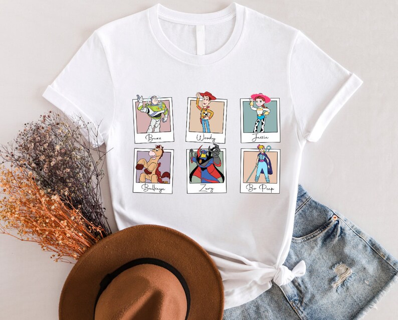 Puede incluir: Camiseta blanca con un gr&aacute;fico de seis personajes de Toy Story en marcos Polaroid. Los personajes son Buzz Lightyear, Woody, Jessie, Bullseye, Zurg y Bo Peep.