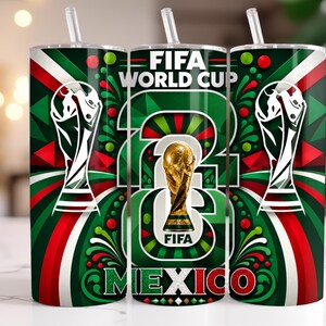 May include: A cylindrical tumbler with a straw featuring a FIFA World Cup theme. The design includes the gold FIFA trophy, the text FIFA WORLD CUP, and MEXICO in green, white, and red colours with geometric patterns and swirling accents.