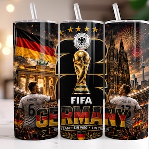 May include: A cylindrical tumbler with a straw featuring a German football theme. The design includes the German flag, the Brandenburg Gate, the Cologne Cathedral, the FIFA World Cup trophy, and players Kimmich and M&uuml;ller. Text includes FIFA, GERMANY, and EIN TEAM EIN WEG EIN TRAUM.