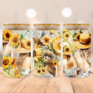 Op de afbeelding: Drie heldere glazen bekers met bamboe deksels en rietjes. Elke beker heeft een aquarelontwerp met zonnebloemen, bladeren en twee vrouwen die strohoeden dragen.