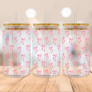 Puede incluir: Tres vasos de vidrio transparentes con patrones de lazos rosas y tapas de bambú. Cada vaso tiene una pajita.