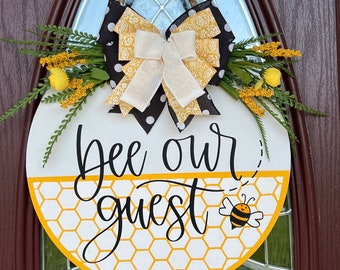 May include: A round wooden door hanger with a yellow honeycomb pattern and the text "Bee our guest" in black script. A black and white polka dot bow with a yellow floral pattern is attached to the top of the hanger. A small cartoon bee is in the bottom right corner.