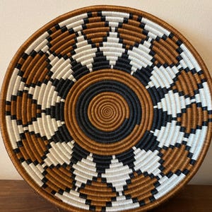 May include: Round woven basket with a spiral center and geometric diamond pattern. The basket features alternating sections of brown, black, and white. The rim and center spiral are brown, creating a visually appealing design. This decorative basket is suitable for home decor.