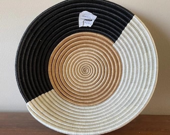 Puede incluir: Una cesta redonda tejida con un diseño en espiral. La cesta está dividida en tres secciones: negro, beige y blanco. La cesta está sobre una superficie de madera.