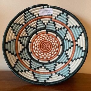 以下が含まれることがあります： 黒、白、青、オレンジの幾何学模様が織り込まれた円形のバスケット。バスケットは深い中心部を持ち、天然素材で作られています。