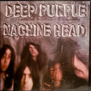 May include: A vintage vinyl record album cover featuring the band Deep Purple. The album title "Machine Head" is embossed in silver lettering on a dark gray background. Three blurry figures are visible in the background.