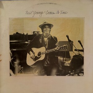 Puede incluir: Portada de álbum en tonos sepia con Neil Young tocando una guitarra acústica. Lleva sombrero y chaqueta. El título del álbum "Comes A Time" está escrito a mano en la parte superior.