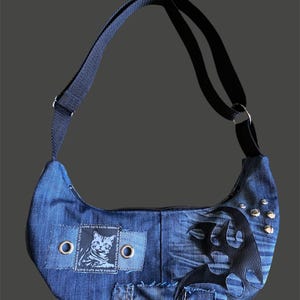 Puede incluir: Bolso de hombro de mezclilla azul con una correa negra y forma de media luna. El bolso presenta un parche con una ilustración de gato y el texto "LOVE CATS HATE MOON". Tiene un diseño de cuero negro y tachuelas plateadas.