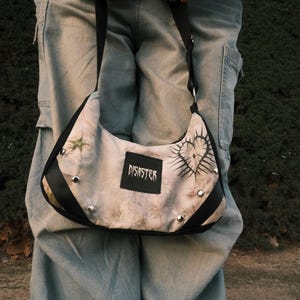 Pode incluir: Uma bolsa de ombro com um design único. A bolsa apresenta um tecido de cor clara com uma estampa, acabamentos pretos e tachas prateadas. A palavra "DISASTER" é impressa em um patch preto.