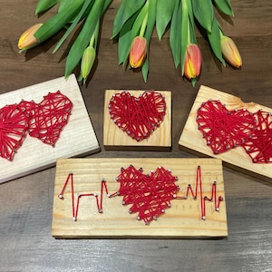 Könnte beinhalten: Vier Holzplaketten mit roten String-Art-Herzen. Eine Plakette zeigt zwei Herzen, eine andere zeigt ein einzelnes Herz und die letzten beiden Plaketten zeigen ein einzelnes Herz mit einer roten String-Art-Herzschlaglinie.