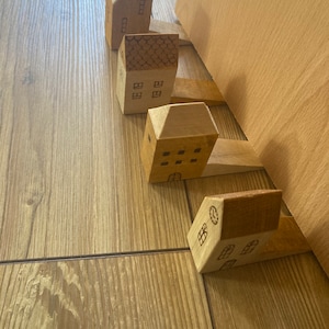 Puede incluir: Cuatro topes de puerta de madera con forma de casa en varios tamaños y diseños. Los topes de puerta están dispuestos en una fila, con el tope de puerta más grande en la parte inferior y el tope de puerta más pequeño en la parte superior. Los topes de puerta están hechos de madera marrón claro y tienen un acabado natural.