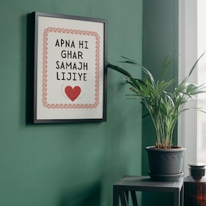 May include: A framed print with a black frame and a white background. The print features a red heart with a stitched design and the text "APNA HI GHAR SAMAJH LIJIYE" in black.