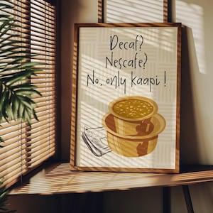 May include: A framed print with the text "Decaf? Nescafe? No, only kaapi!" features a drawing of two stacked coffee cups and a newspaper. The artwork is in a wooden frame, set against a beige patterned background.