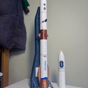 May include: A model rocket with a white and copper body, featuring the NASA logo and the text "NEW GLENN" and "BLUE ORIGIN". The rocket is displayed on a blue stand, with a smaller white rocket beside it.
