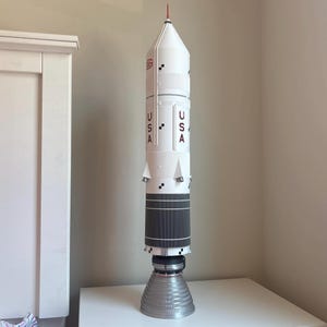 May include: A tall, white model rocket featuring a red, pointed tip, dark gray cylindrical sections, and a large silver engine nozzle at the base. The body displays the text USA in red lettering, along with small black square markings and structural details.