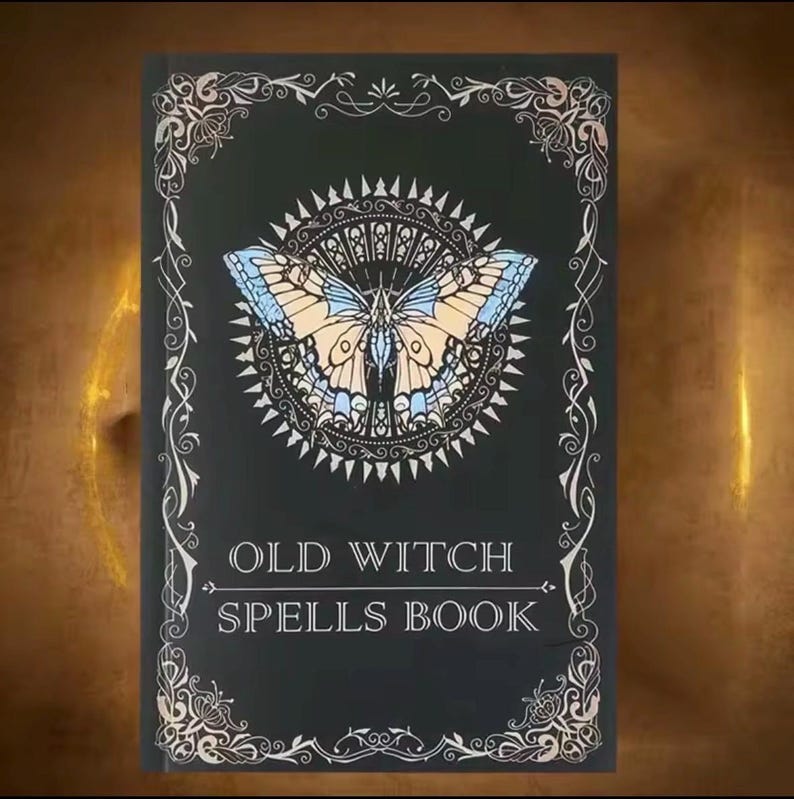 以下が含まれることがあります： 「OLD WITCH SPELLS BOOK」のテキストが入った黒い本の表紙。青とベージュの蝶のイラストが特徴で、装飾的なサンバーストのデザインで囲まれています。装飾的な銀色のスクロールワークが縁を縁取っています。